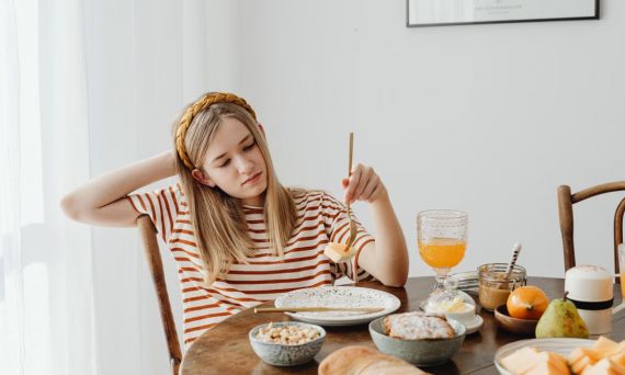Teen girl in striped shirt at breakfast table with fruits, bread, and juice. Casual morning setting.